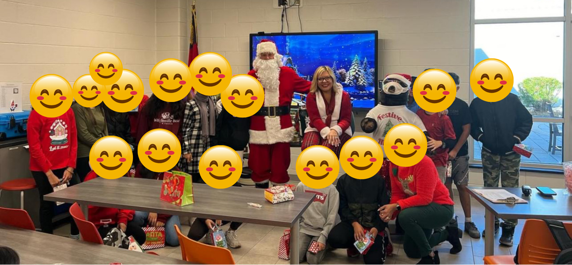 Group of children and adults celebrating Christmas with Santa Claus in a room decorated for the holidays. Everyone is smiling and holding gift bags or presents, with a large illustrated Christmas scene on the TV screen behind them.