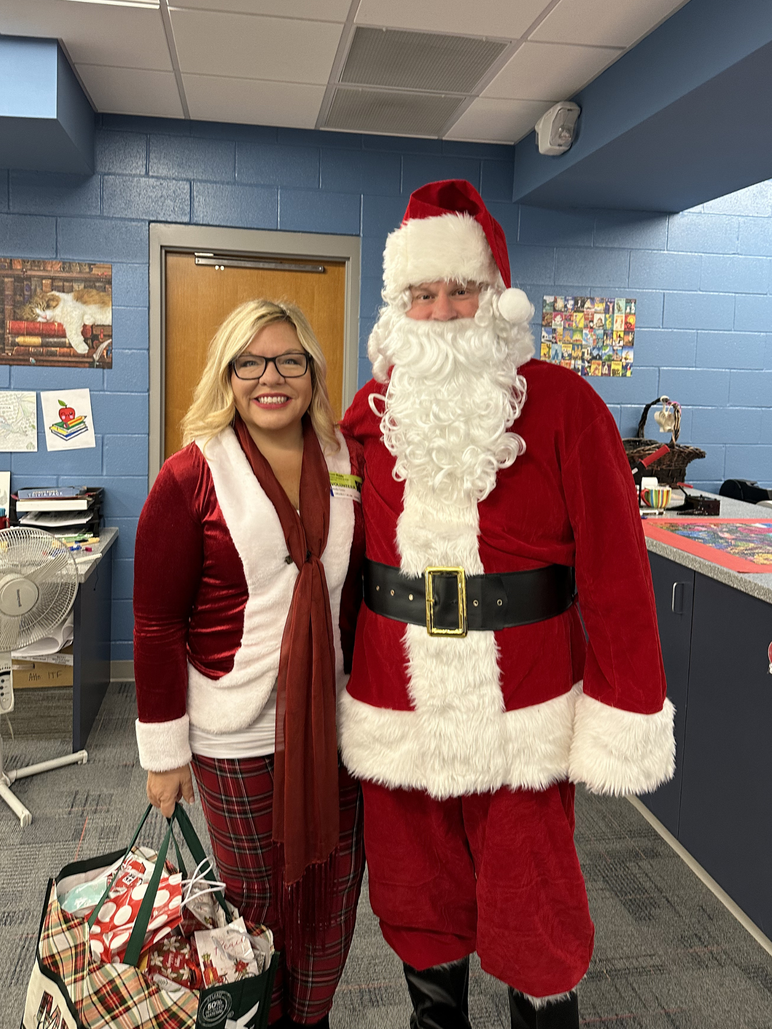 A woman dressed in holiday plaid pants, a red and white shirt, and black glasses stands next to a man dressed as Santa Claus. They are indoors in a room with blue walls decorated with children's artwork, and holiday decorations in the background.