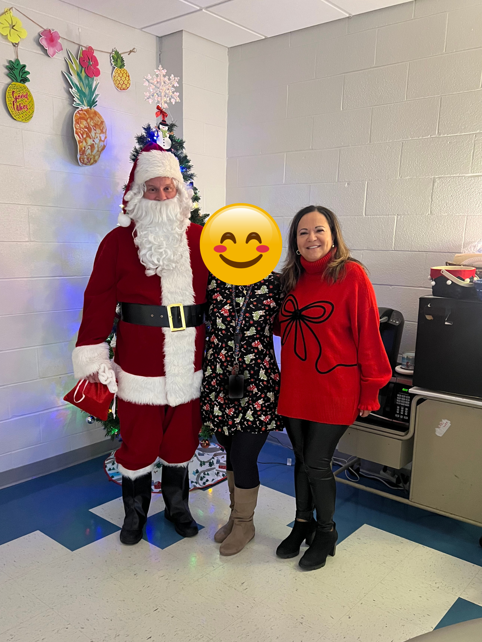 Three people standing together, with a decorated Christmas tree in the background. The person on the left is dressed as Santa Claus, the person in the middle has a holiday-themed face emoji, and the person on the right is wearing a red sweater with a black bow design.