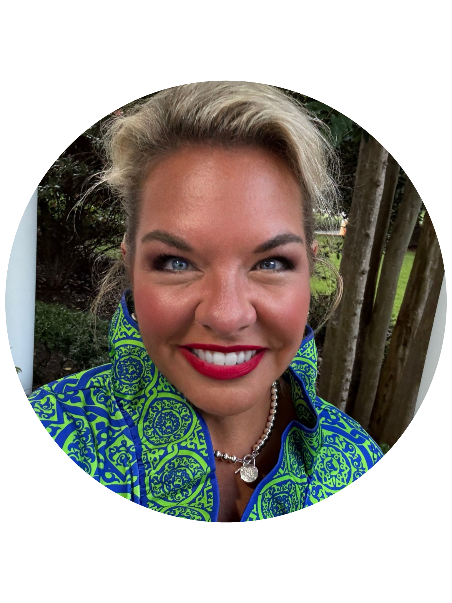 A woman with blonde hair and blue eyes smiling outdoors in front of a wooden fence and greenery, wearing a bright blue and green patterned jacket and a pearl necklace.