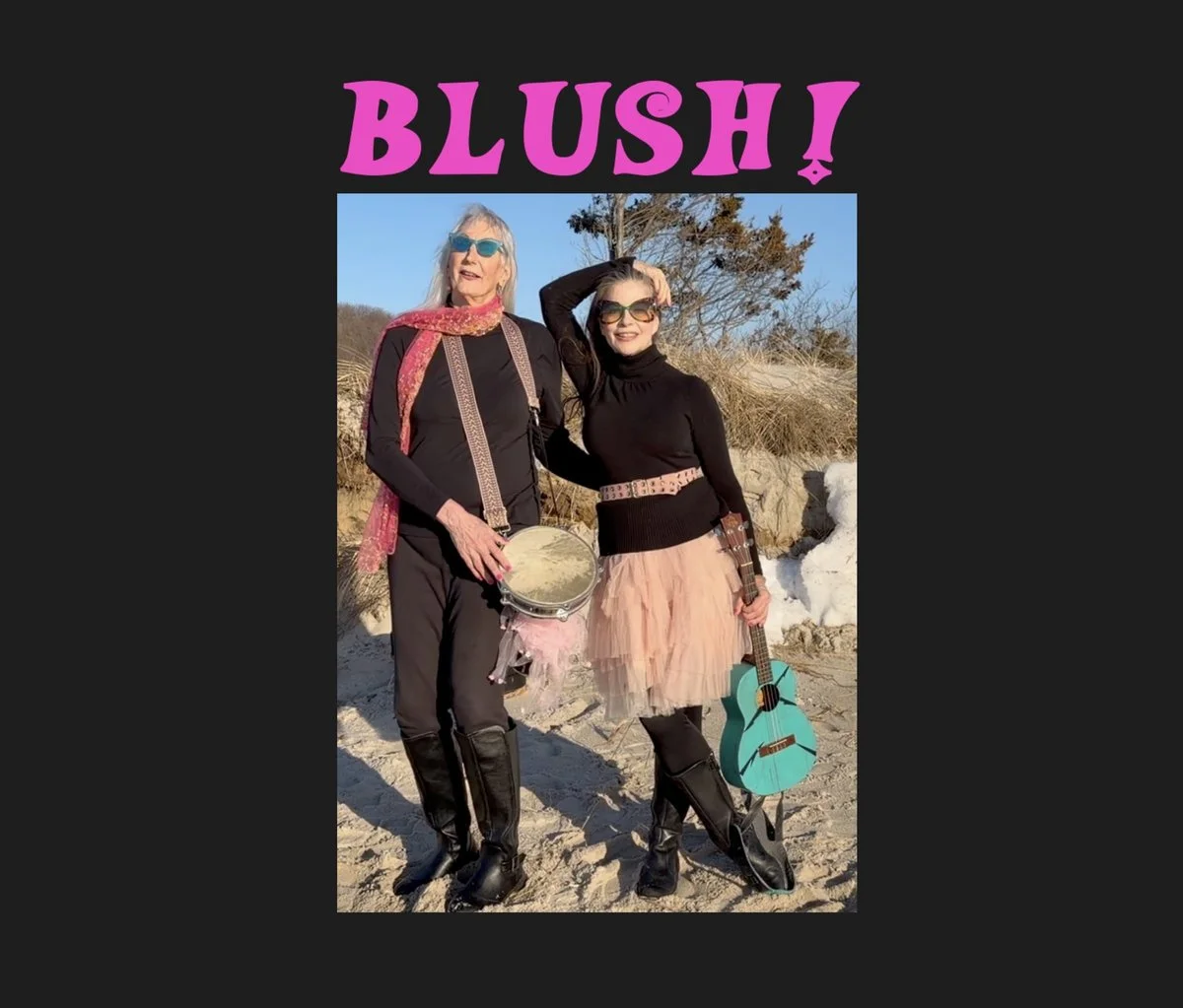 Two women standing on a sandy beach outdoors, smiling and dressed in black outfits with accessories. One is holding a tambourine, the other has a turquoise guitar. The background features sand dunes, some snow, leafless trees, and a clear blue sky. The word 'BLUSH!' is written in large pink letters at the top of the image.