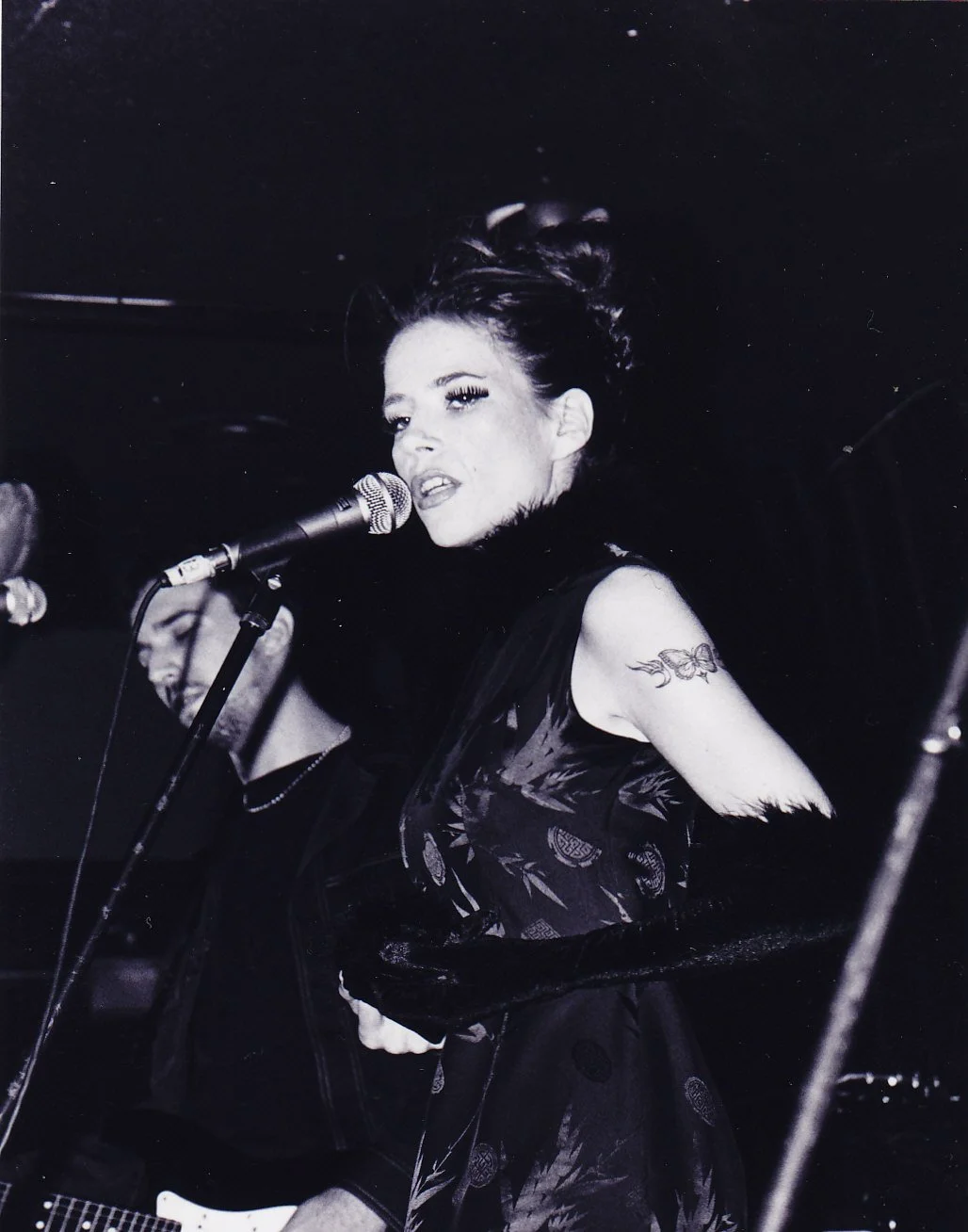 Black and white photo of a woman singing into a microphone with her eyes closed. She has dark hair styled in an updo, prominent eyelashes, and a tattoo on her upper arm. She is wearing a dark dress with a floral or patterned design.