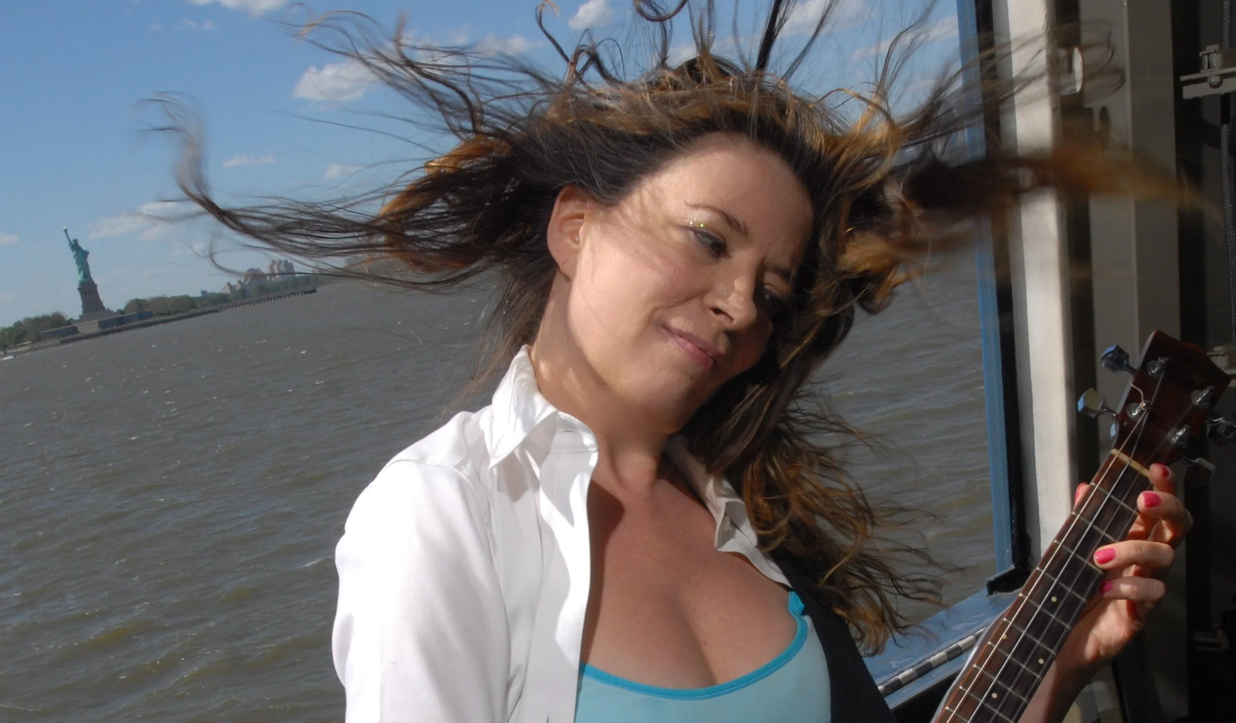Woman with brown hair playing guitar on boat with Statue of Liberty in the background.
