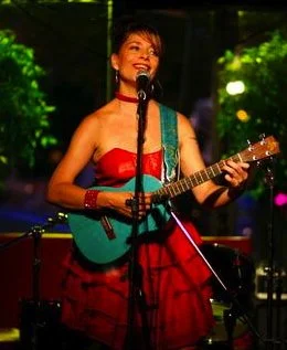 A woman with short dark hair playing a turquoise guitar and singing into a microphone on stage, wearing a red strapless dress and red accessories with green foliage in the background.