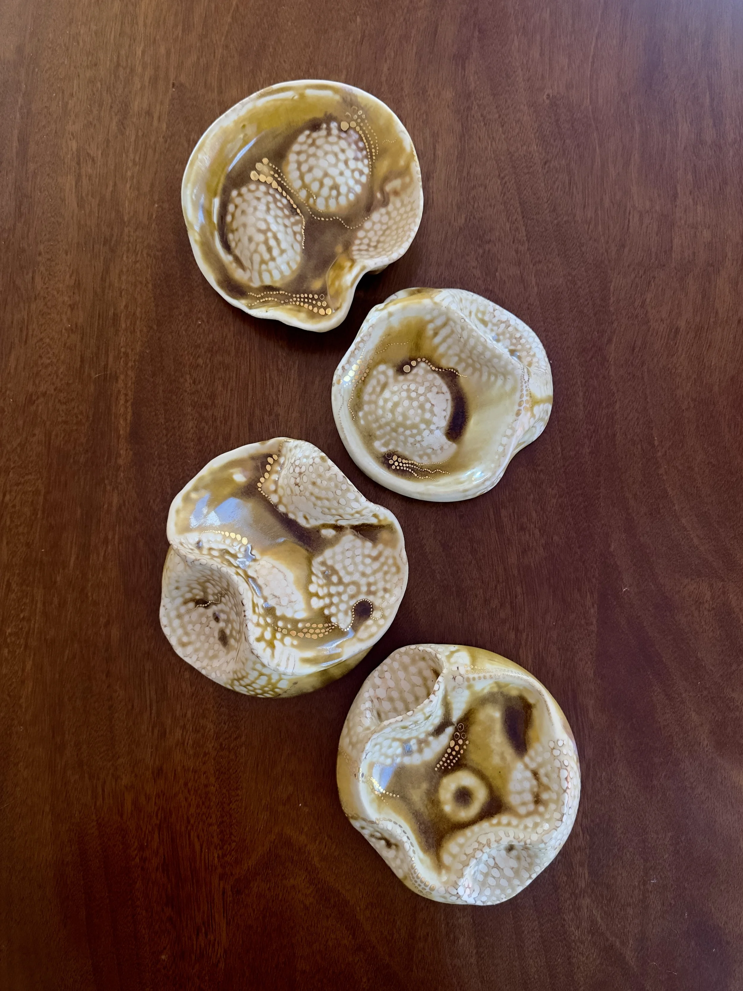 Four circle sculptures with fluid topographical shapes. They are brownish yellow in color and have white dots on them of bare clay. The parts with glaze feel smooth and glassy, and the parts without glaze feel like the unglazed bottom of a coffee mug