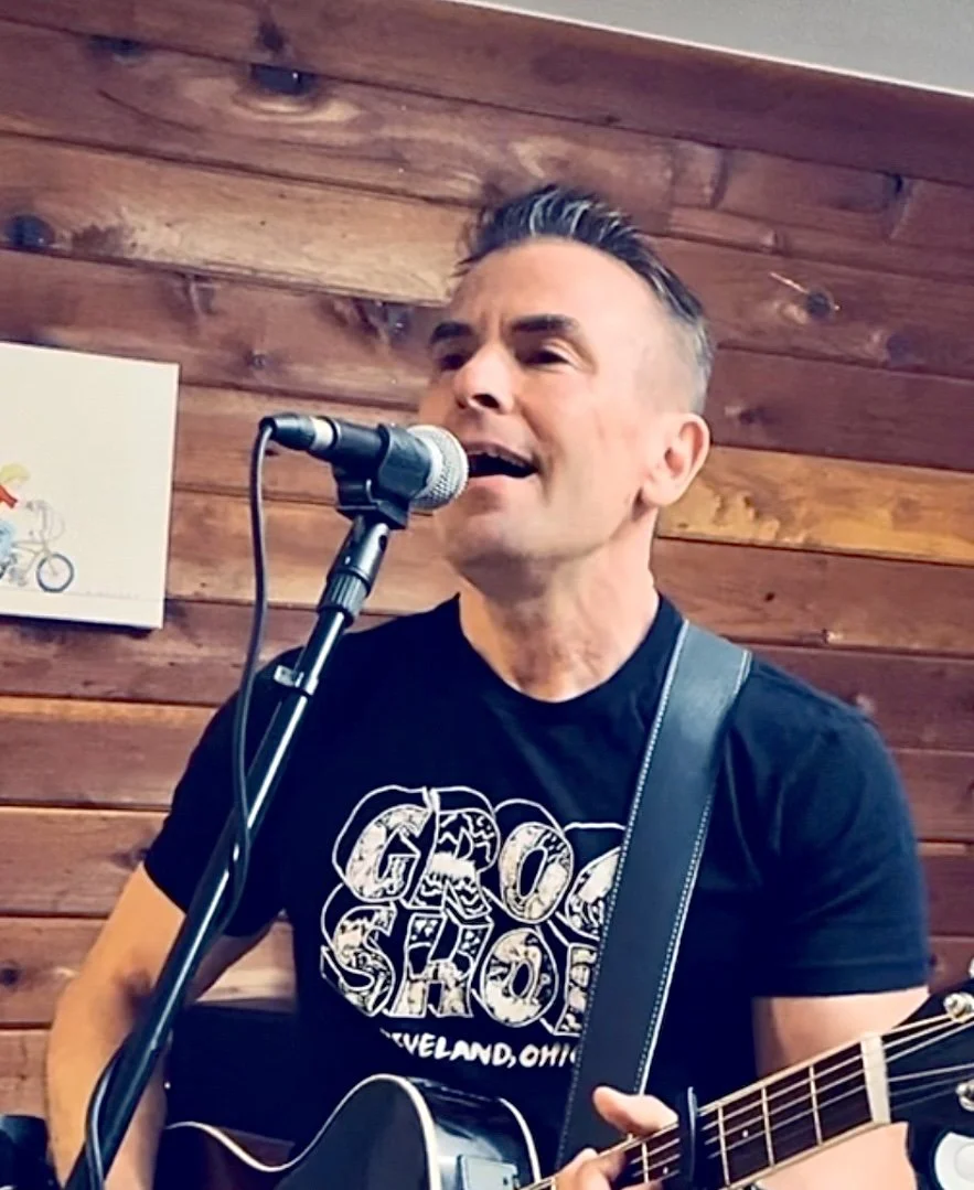 Man singing into a microphone and playing an acoustic guitar in front of a wooden wall, wearing a black T-shirt with white graphic text.