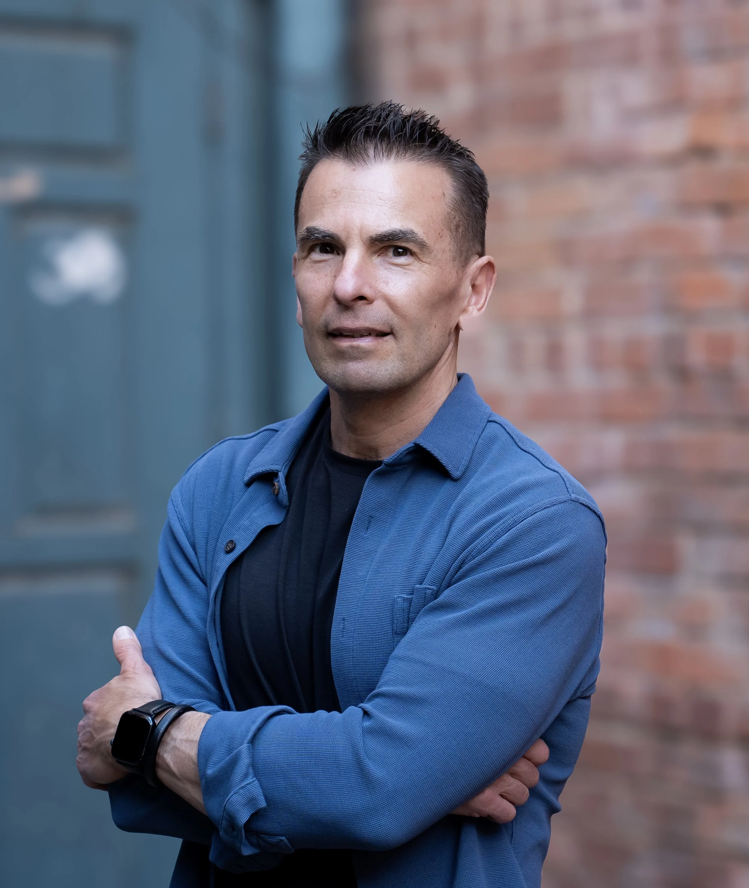 A man with dark, spiked hair, wearing a blue collared shirt and a black T-shirt, crossing his arms in front of a brick wall and a blue door.