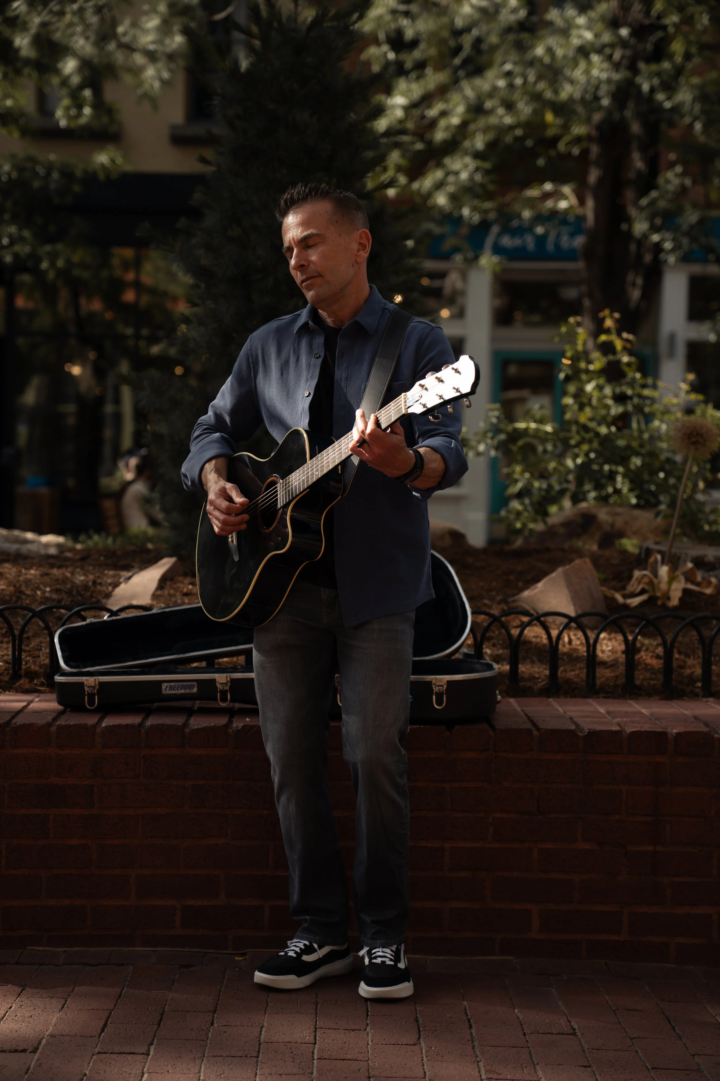 A man playing an acoustic guitar outdoors during the daytime, standing on brick pavement in front of a low brick wall with a guitar case open behind him. He wears a dark blue button-up shirt, black and white sneakers, and has a focused expression on 