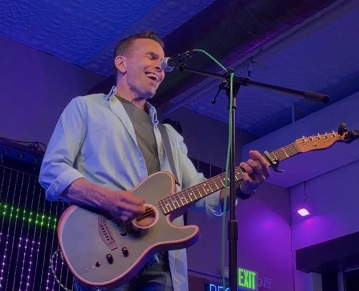A man singing and playing an electric guitar on stage during a live music performance with colorful lighting.
