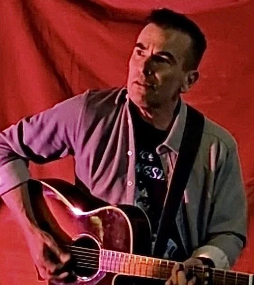 A man playing an acoustic guitar on stage with a red curtain background.