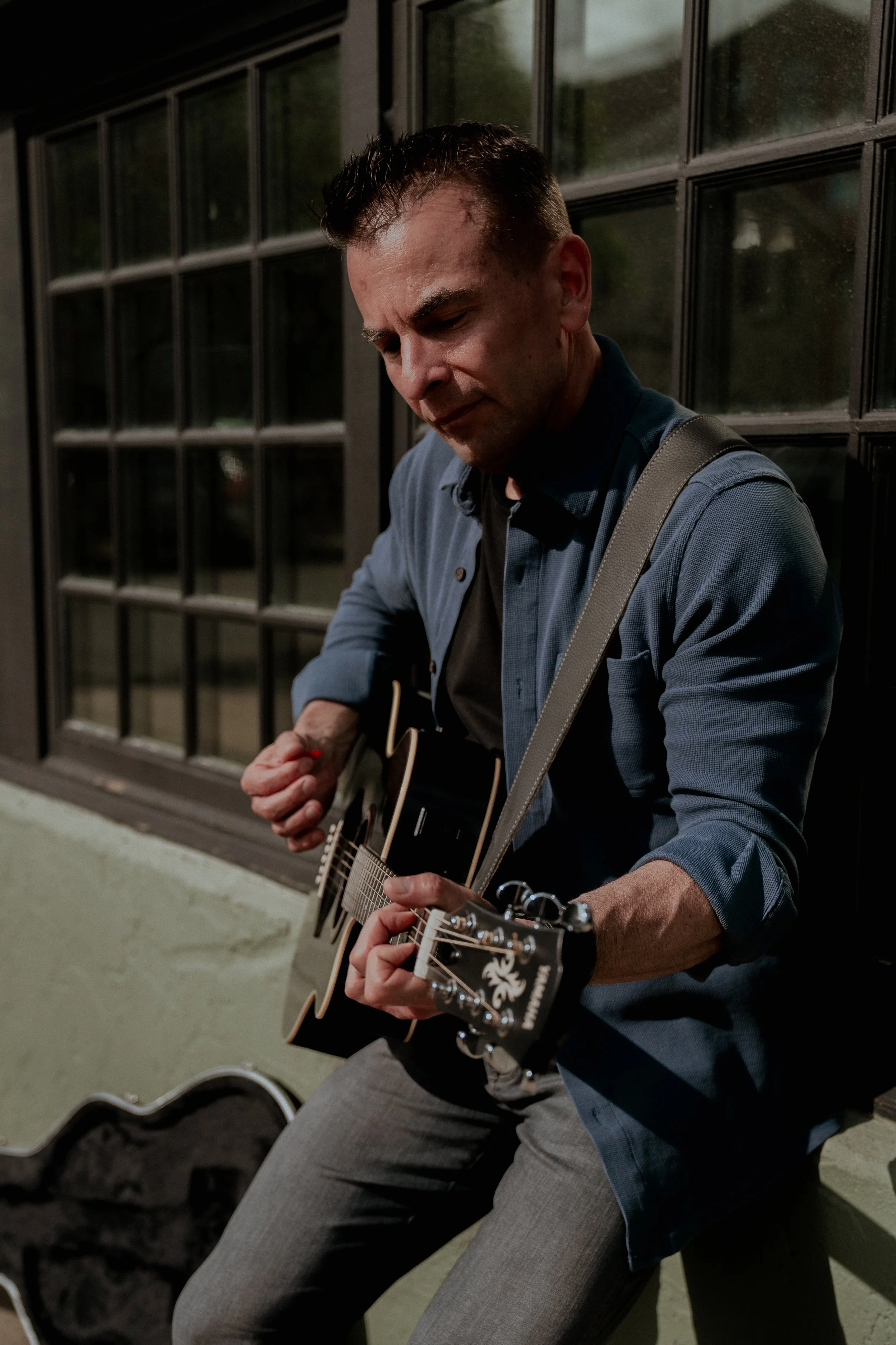 A man with short dark hair, dressed in a blue blazer and gray pants, playing an acoustic guitar with a black and white design on the headstock, sitting against a black grid window, with a guitar case on the ground nearby.