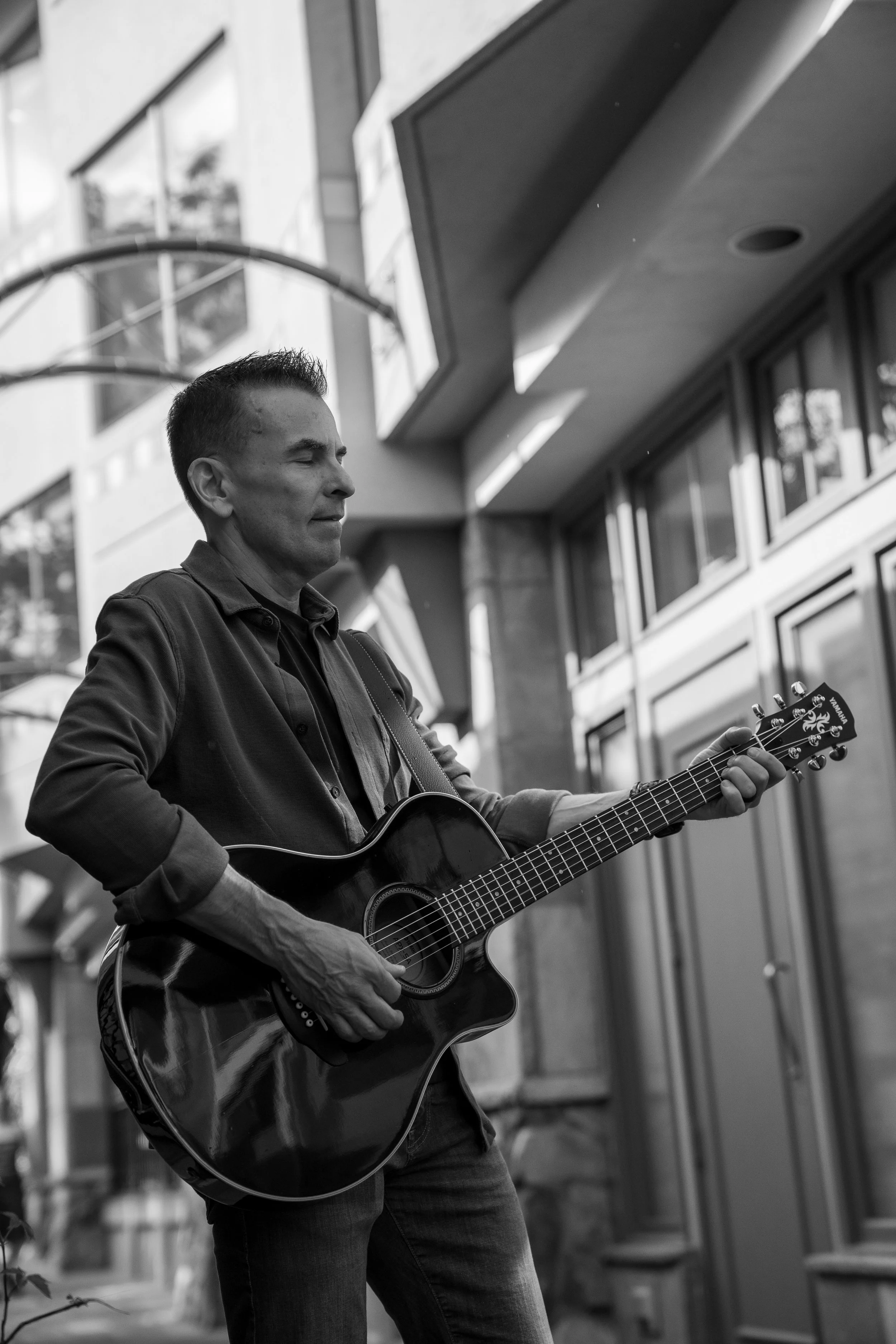 A man playing an acoustic guitar outdoors in an urban setting, with modern buildings and large windows in the background.