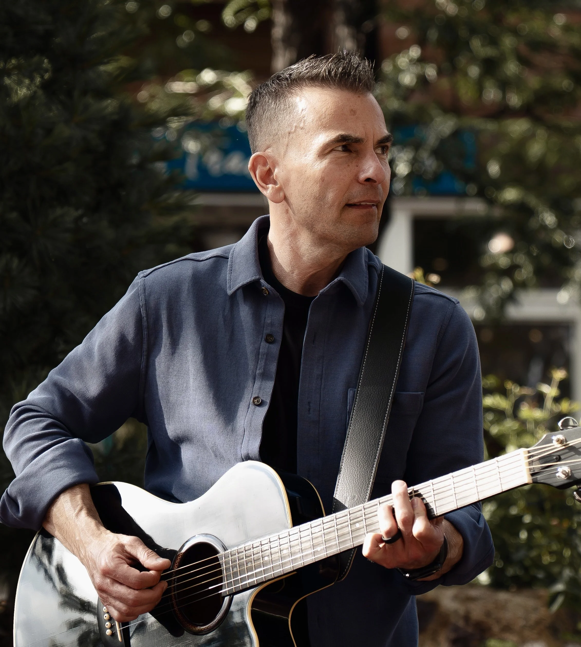 Man playing an acoustic guitar outdoors, wearing a dark blue shirt, with trees and blurred background.