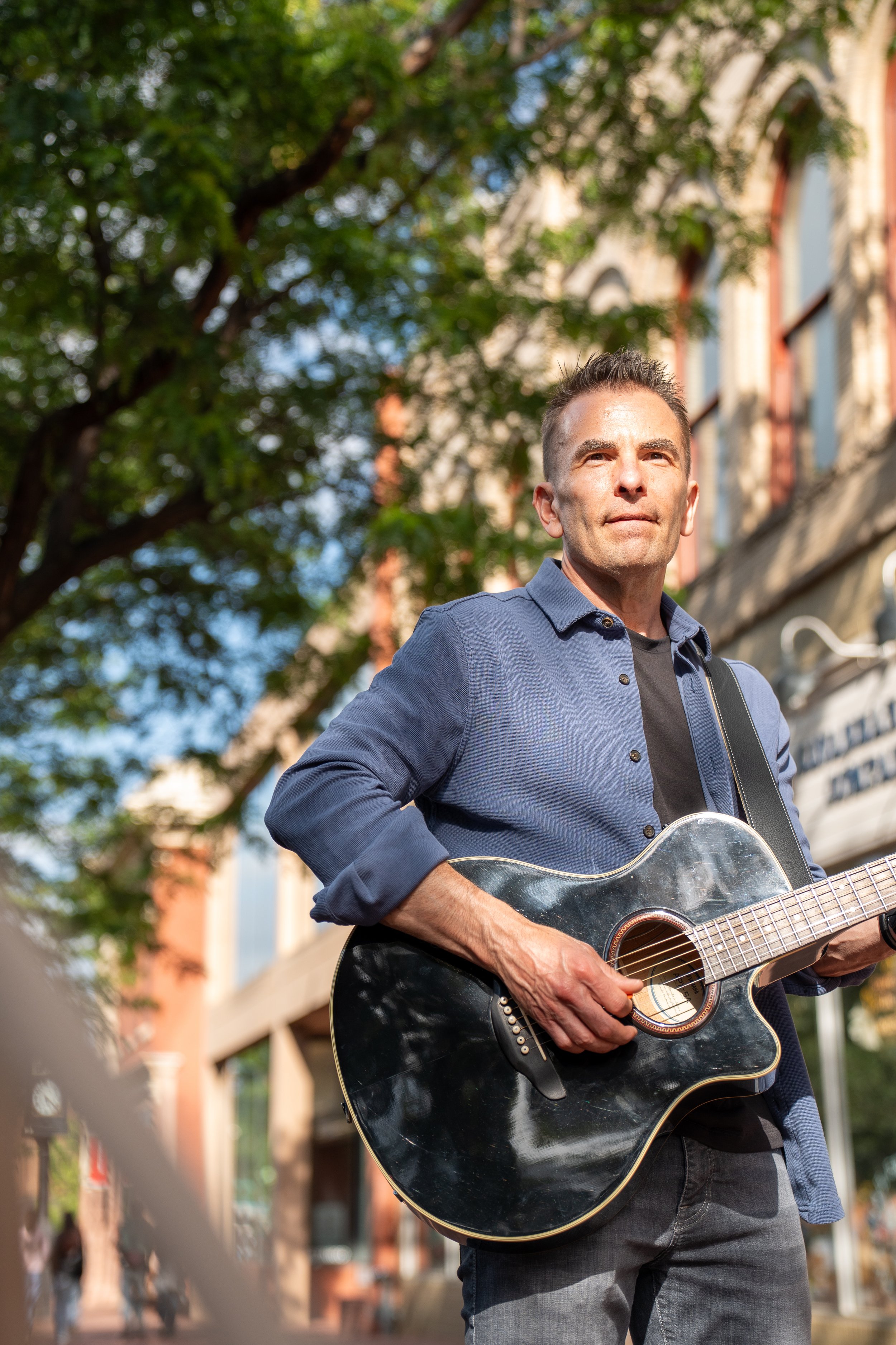 Man playing black acoustic guitar outdoors in city with trees and buildings.