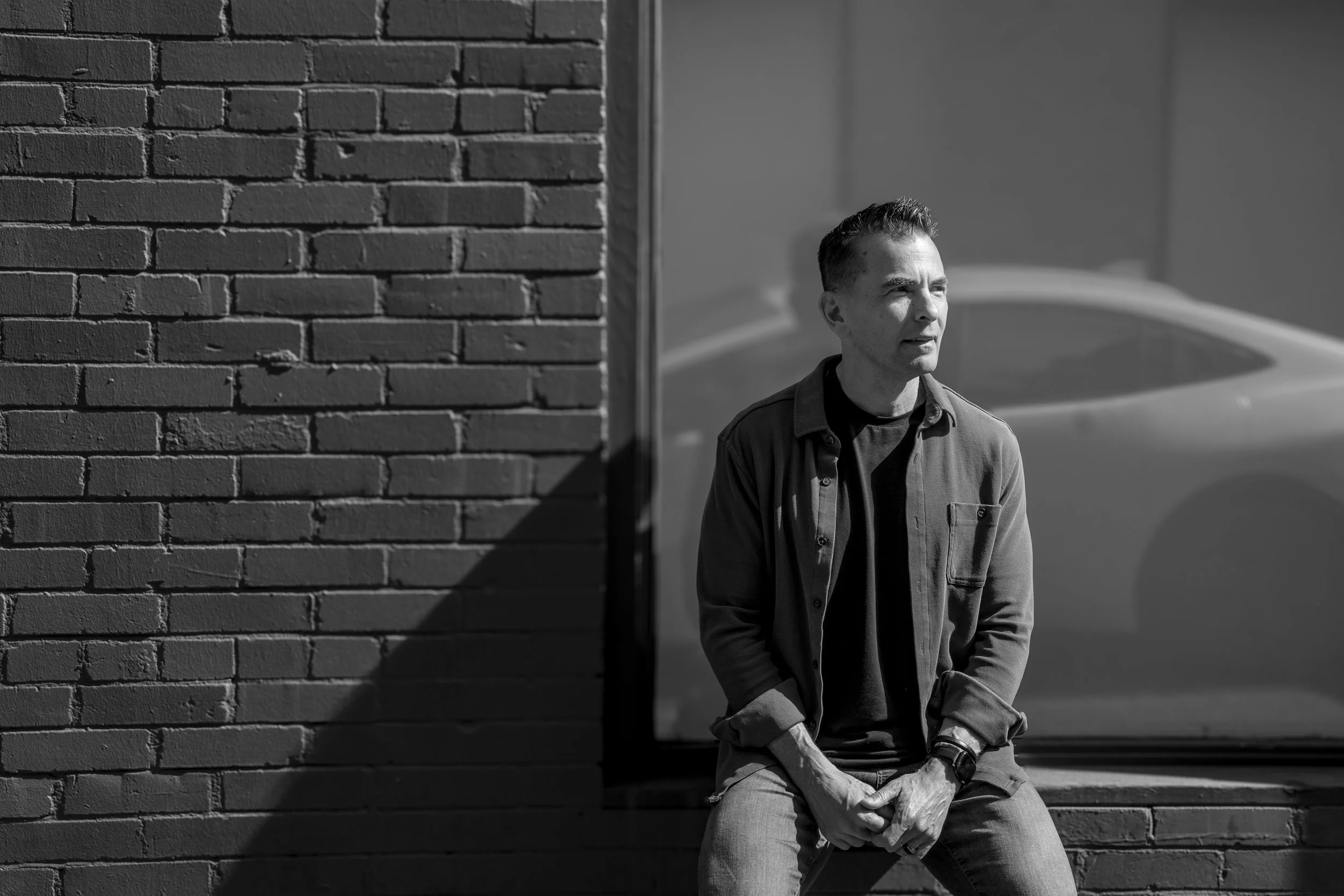 A man sitting on a ledge next to a brick wall, with a car and building in the background, in black and white.