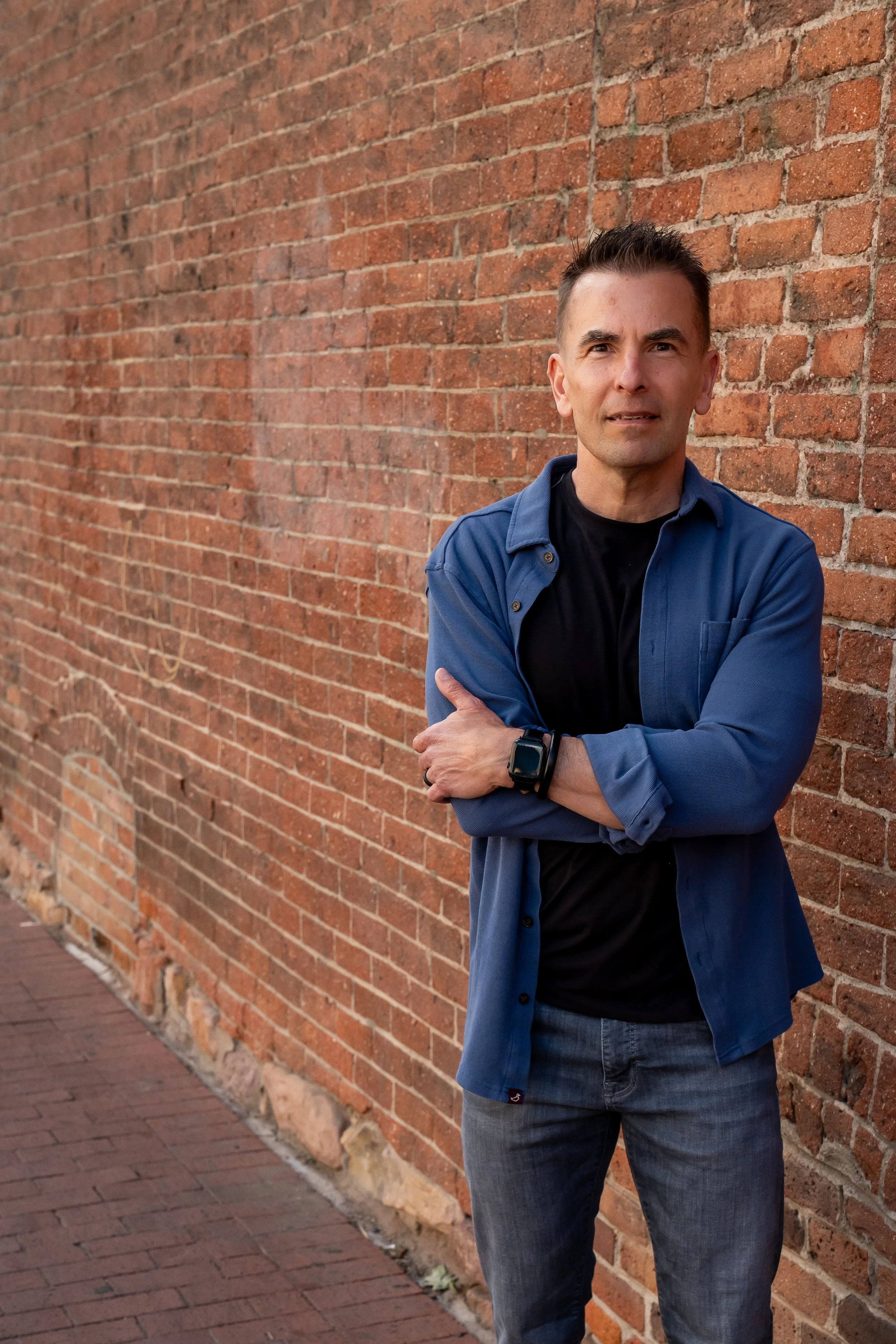 A man with short dark hair, wearing a black t-shirt, a blue button-up shirt, and jeans, standing with arms crossed against a red brick wall.