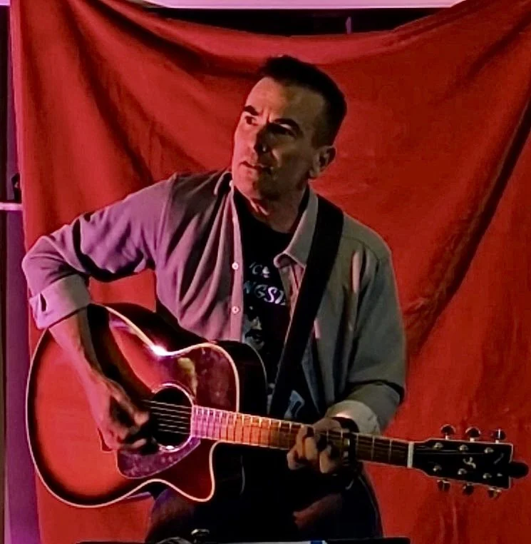 A man playing an acoustic guitar with a red background behind him.