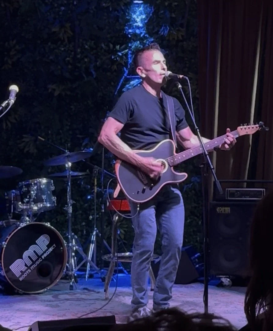 Male musician playing an acoustic guitar and singing into a microphone on stage with a drum set in the background, during a live performance.