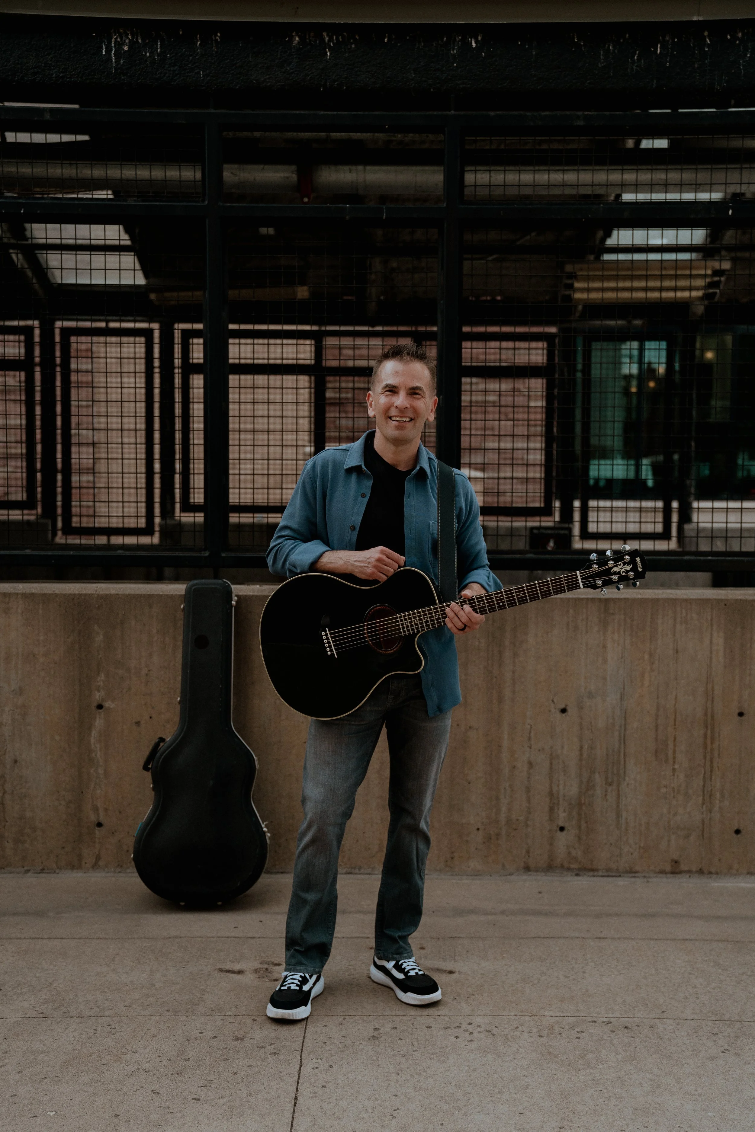 A man standing outdoors with a black acoustic guitar in a black case beside him, smiling, wearing a blue jacket over a black shirt, gray jeans, and black sneakers with white soles. Behind him is a metal fence structure with brick walls and windows, a