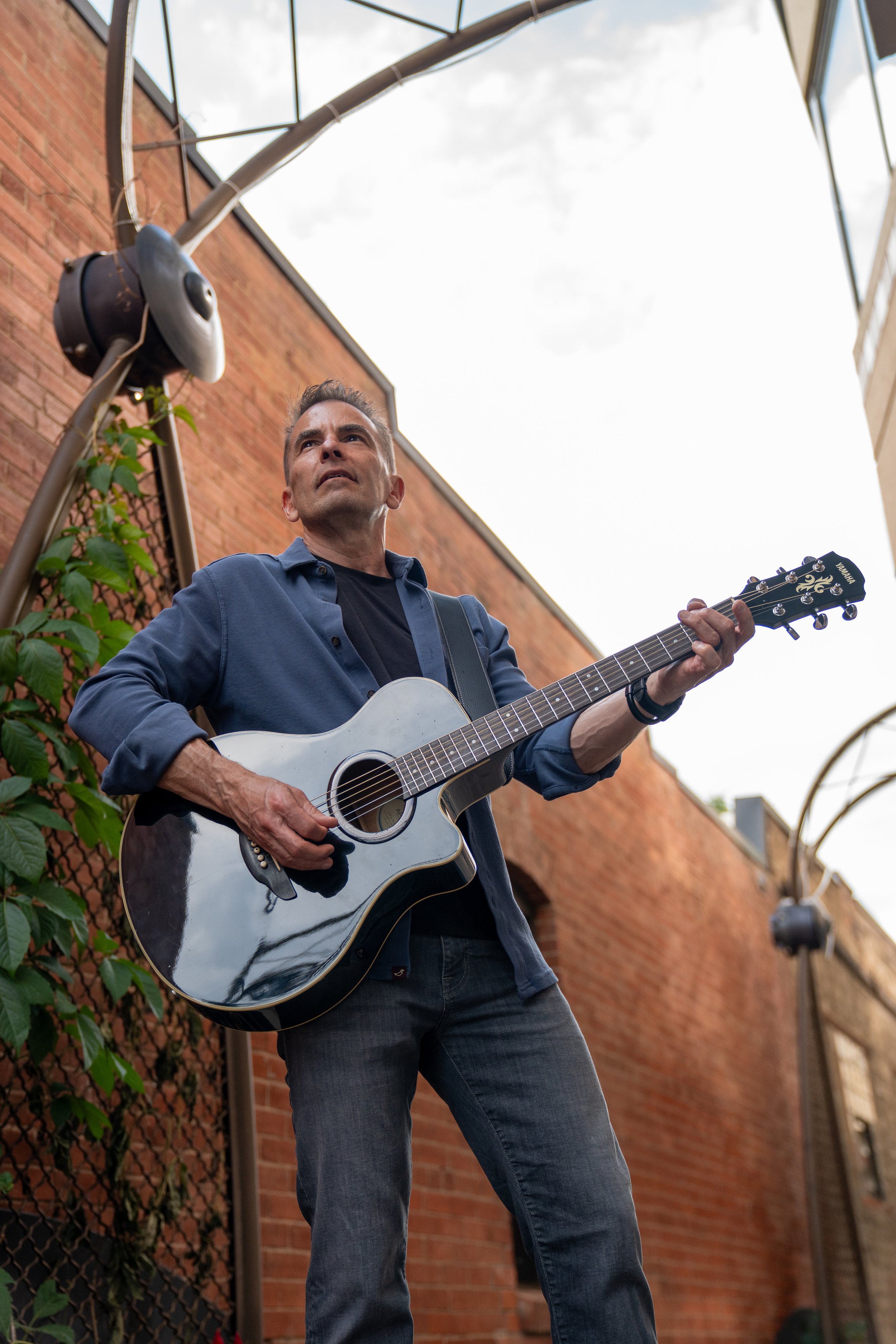 A man playing an acoustic guitar outdoors on a narrow alley with brick walls and glass at the top, looking upwards with a focused expression.