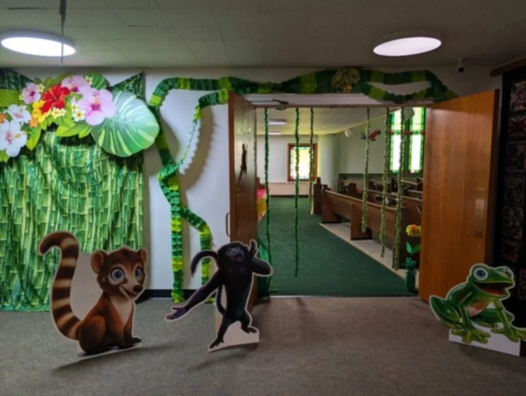 Decorative jungle scene with cartoon animals including a lemur, a tiger, and a frog, leading into a room with wooden benches and green streamers hanging from the ceiling.