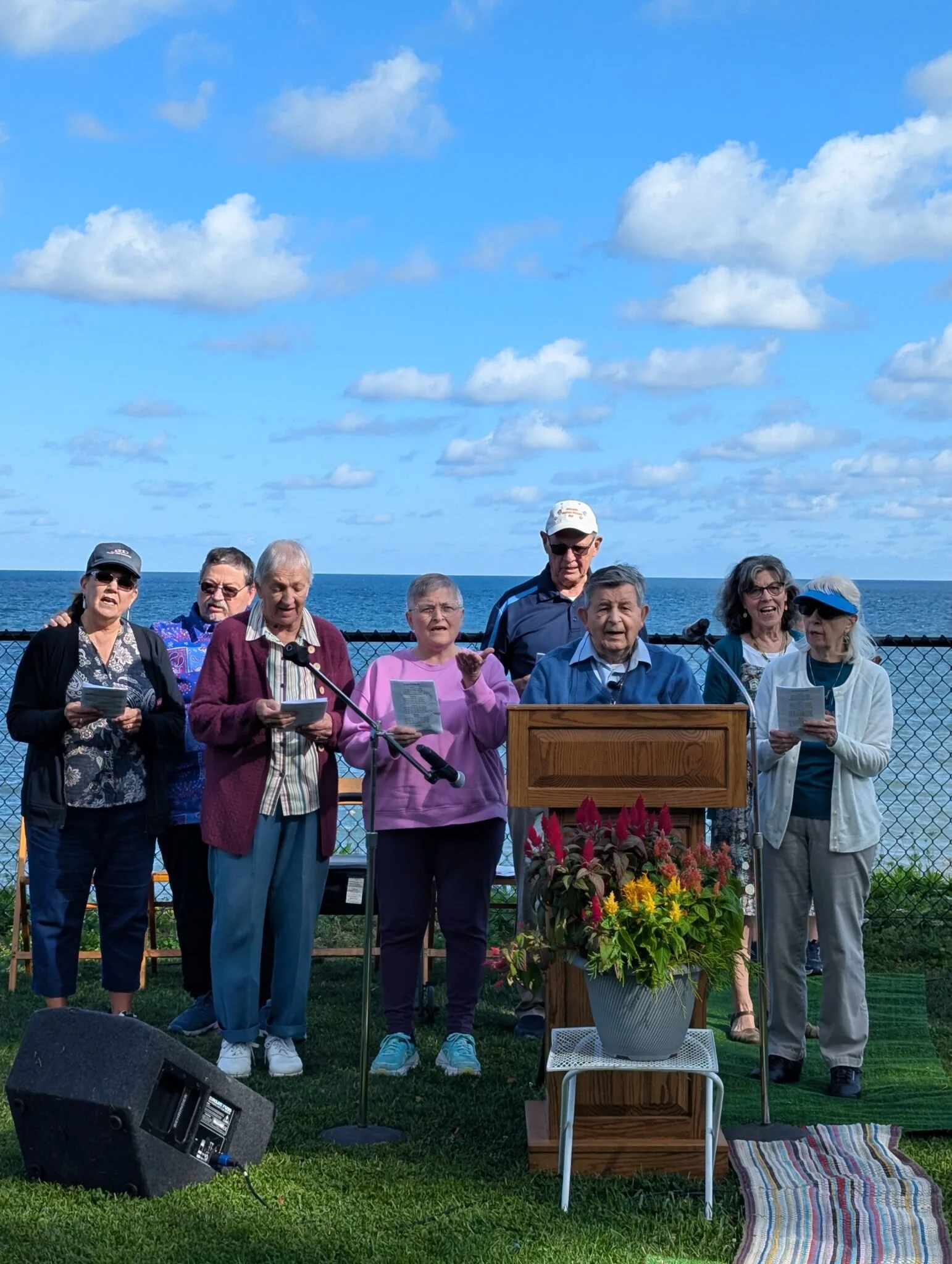 A group of people singing or speaking outdoors near the ocean, with a podium, flowers, and a microphone, under a partly cloudy sky.