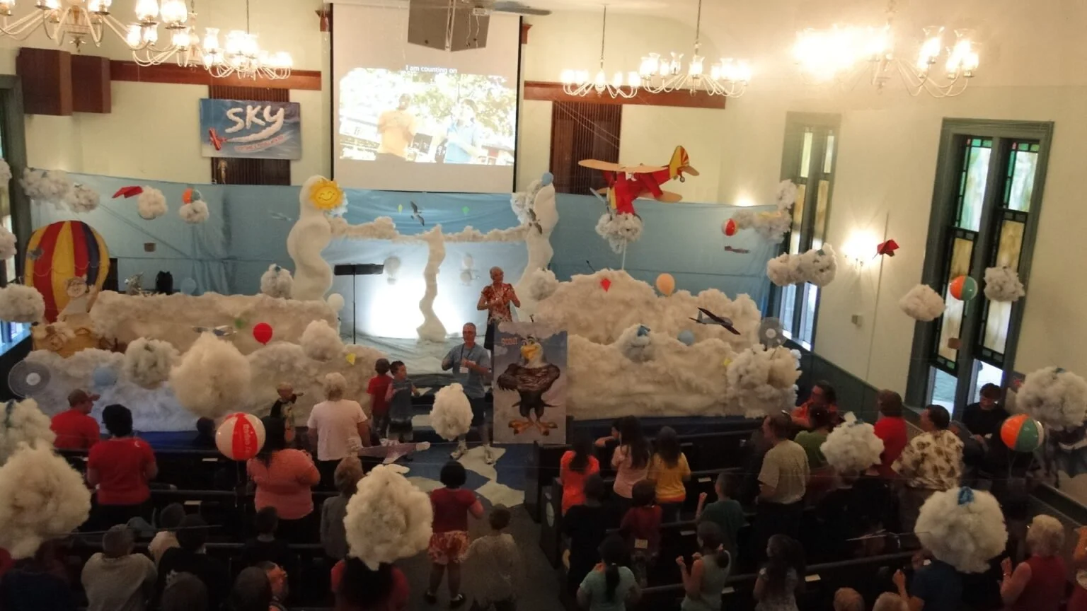 Children and adults in an auditorium watching a stage decorated with clouds, balloons, and a flying airplane, with a large screen above displaying a video.