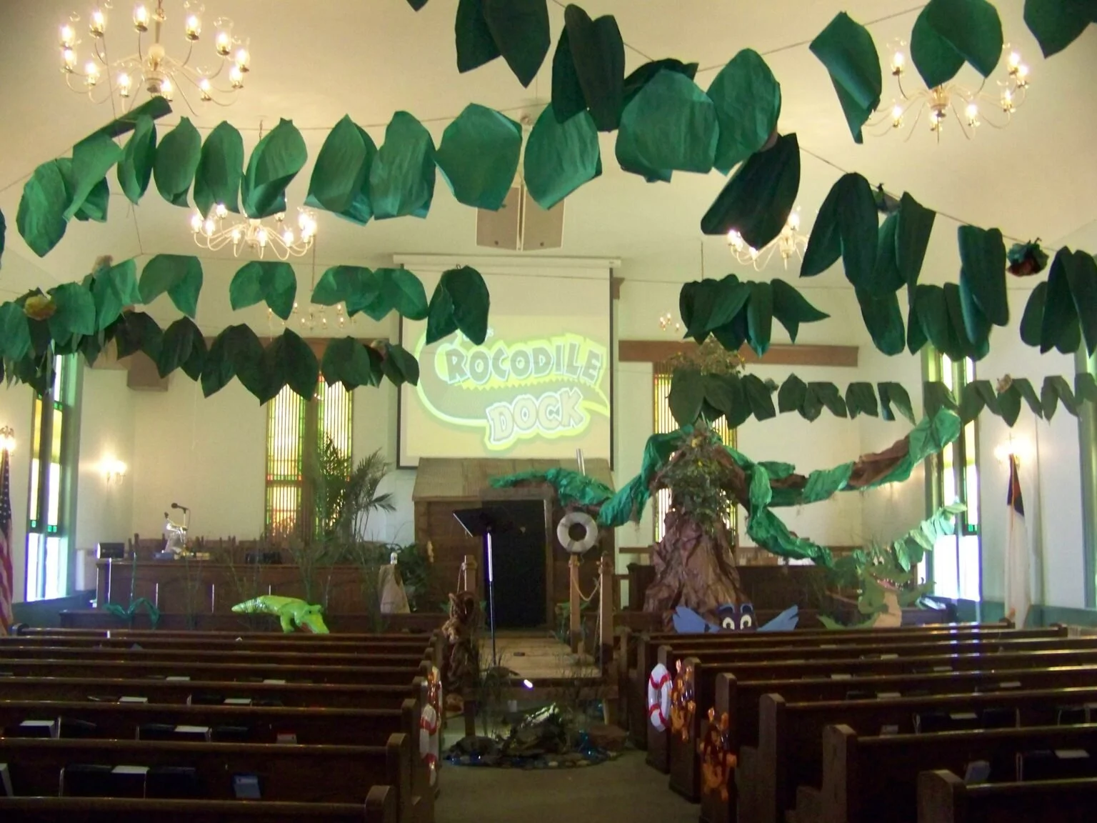 Decorated church interior with jungle theme, featuring large green leaves hanging from the ceiling, a large painted tree prop, a monitor with 'Crocodile Dock' in bright yellow letters, and small stuffed crocodile toys.