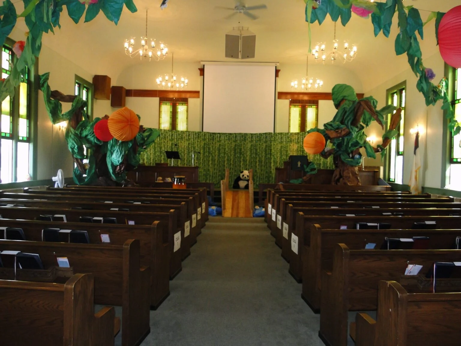 Inside a church decorated with paper trees and lanterns, with a panda plush on the stage, and chairs arranged for a gathering.
