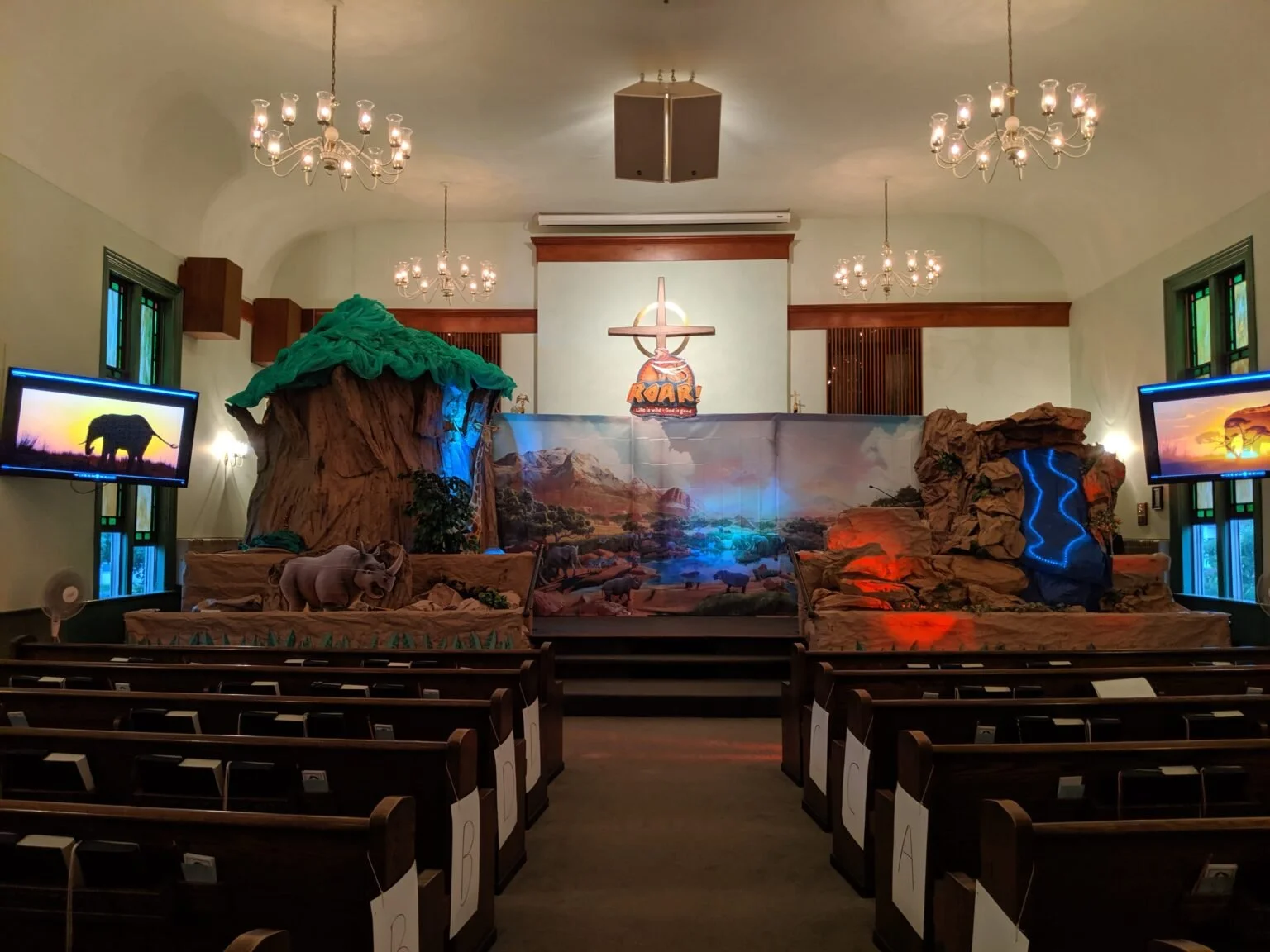 Stage set for a biblical play with desert landscape backdrop, cardboard mountains, and animal props, including a lion and waterfall, in a church with chandeliers and stained glass windows.
