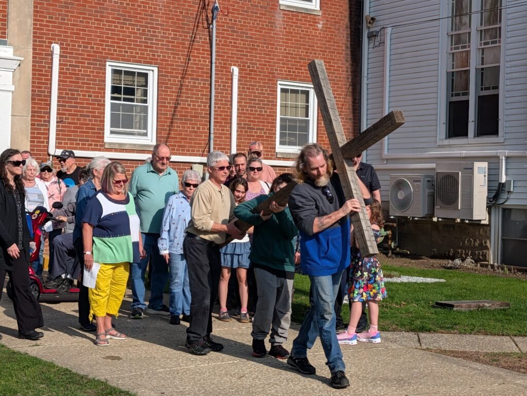 A group of people participating in a religious procession, carrying a large wooden cross through a neighborhood street.