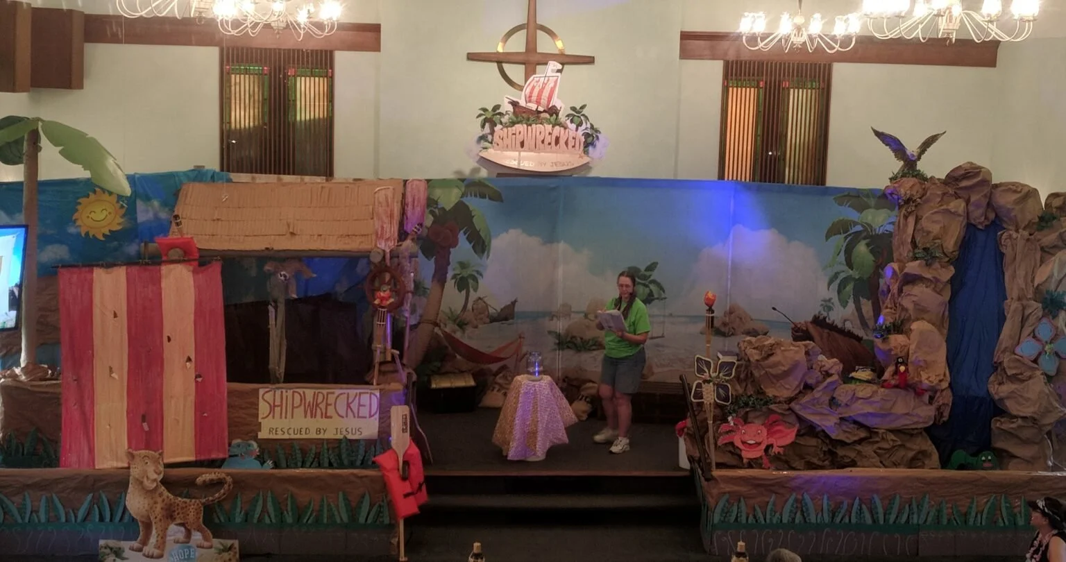 Indoor stage decorated as a tropical island with a painted mural, palm trees, rocks, and a waterfall. A woman in a green shirt and shorts stands on the stage holding a Bible or book. There is a sign that reads 'SHIPWRECKED RESCUED BY JESUS'. The stage has various decorations including a lion figure, plants, and a backdrop depicting an island. Chandeliers hang from the ceiling.