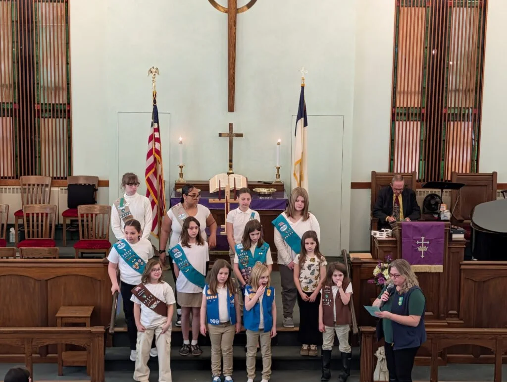 A group of children and a woman in a church, some children wearing Boy Scout vests, standing on the steps in front of the altar. The church interior features wooden accents, American and church flags, and a cross on the wall.