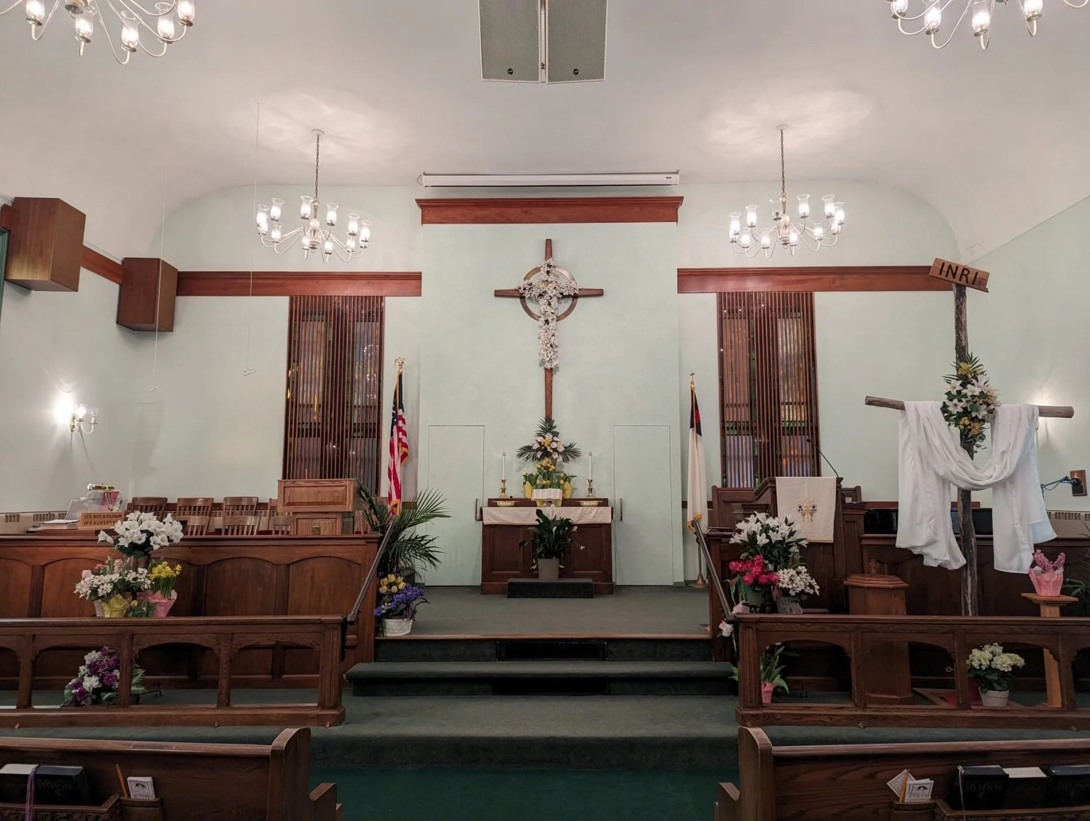 Interior of a church altar decorated with flowers, a crucifix, and American flags, with wooden pews and chandeliers.