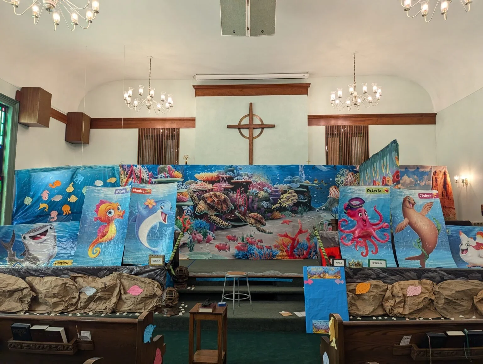 Church altar decorated with ocean-themed decorations for a children's event, featuring colorful images of sea creatures like fish, octopus, turtle, dolphin, seal, and seahorse, with a backdrop of coral and ocean scenes.