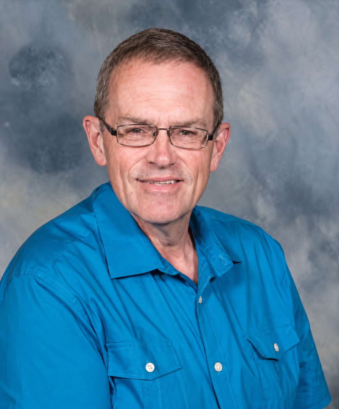 Portrait of a senior-aged man with glasses and short, light brown hair, wearing a bright blue shirt, set against a cloudy gray background.