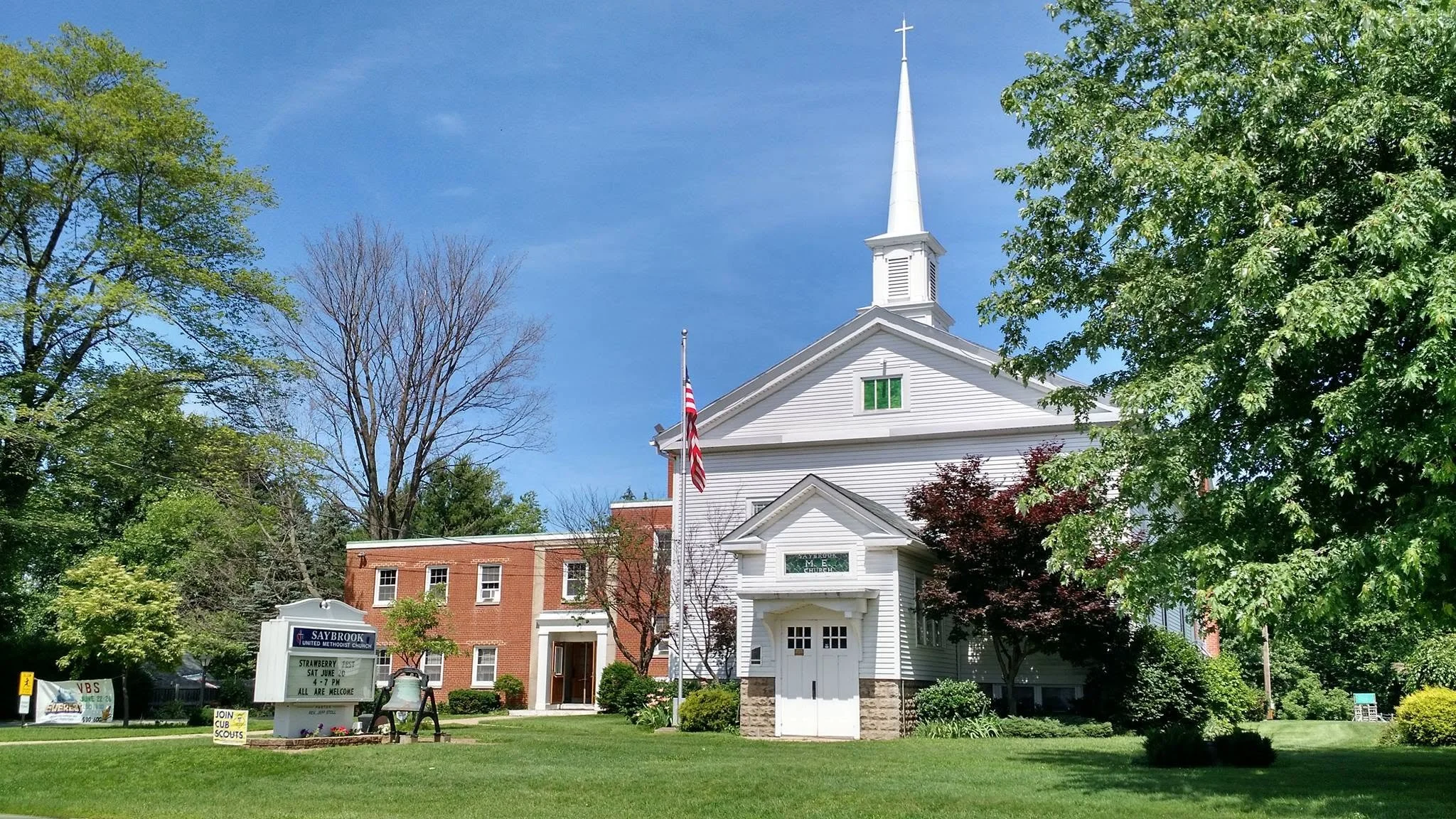 A white church with a tall steeple, surrounded by trees and a lawn, with a blue sky in the background.