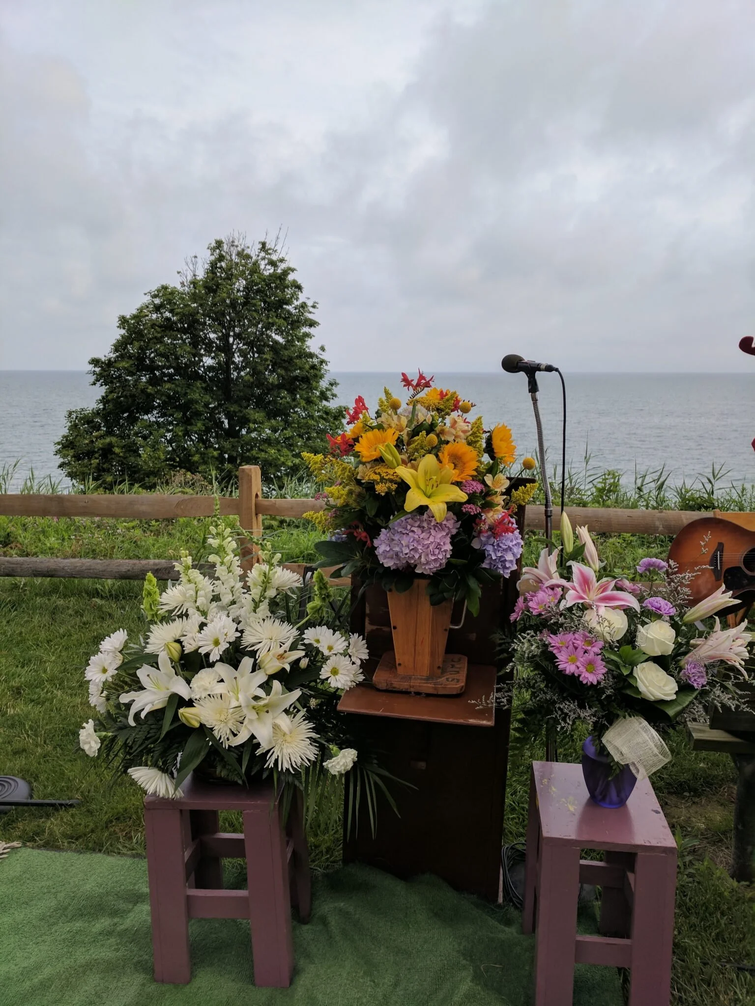 A memorial setup with multiple floral arrangements at an outdoor location by the water, featuring a microphone and a guitar, with a tree and cloudy sky in the background.