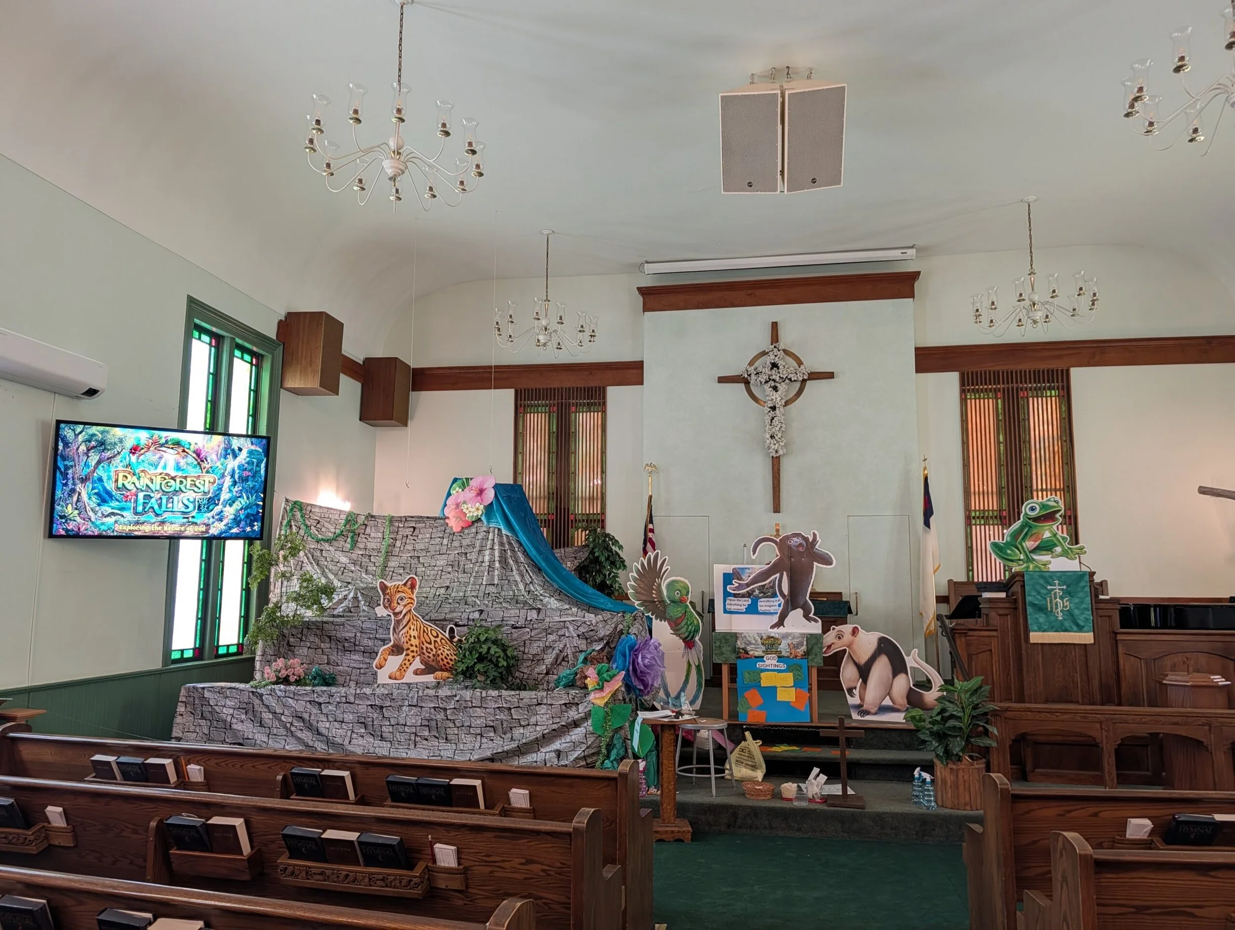 Interior of a church decorated with jungle animals and a waterfall theme, including cutouts of a cheetah, a bear, and a bird, along with plants, flowers, and a TV screen displaying the title "Rainforest Falls".