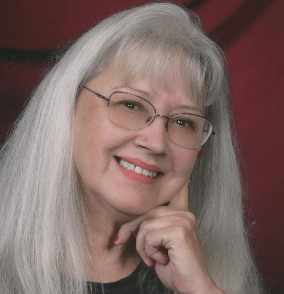 A woman with long gray hair and glasses smiling at the camera, resting her chin on her hand.