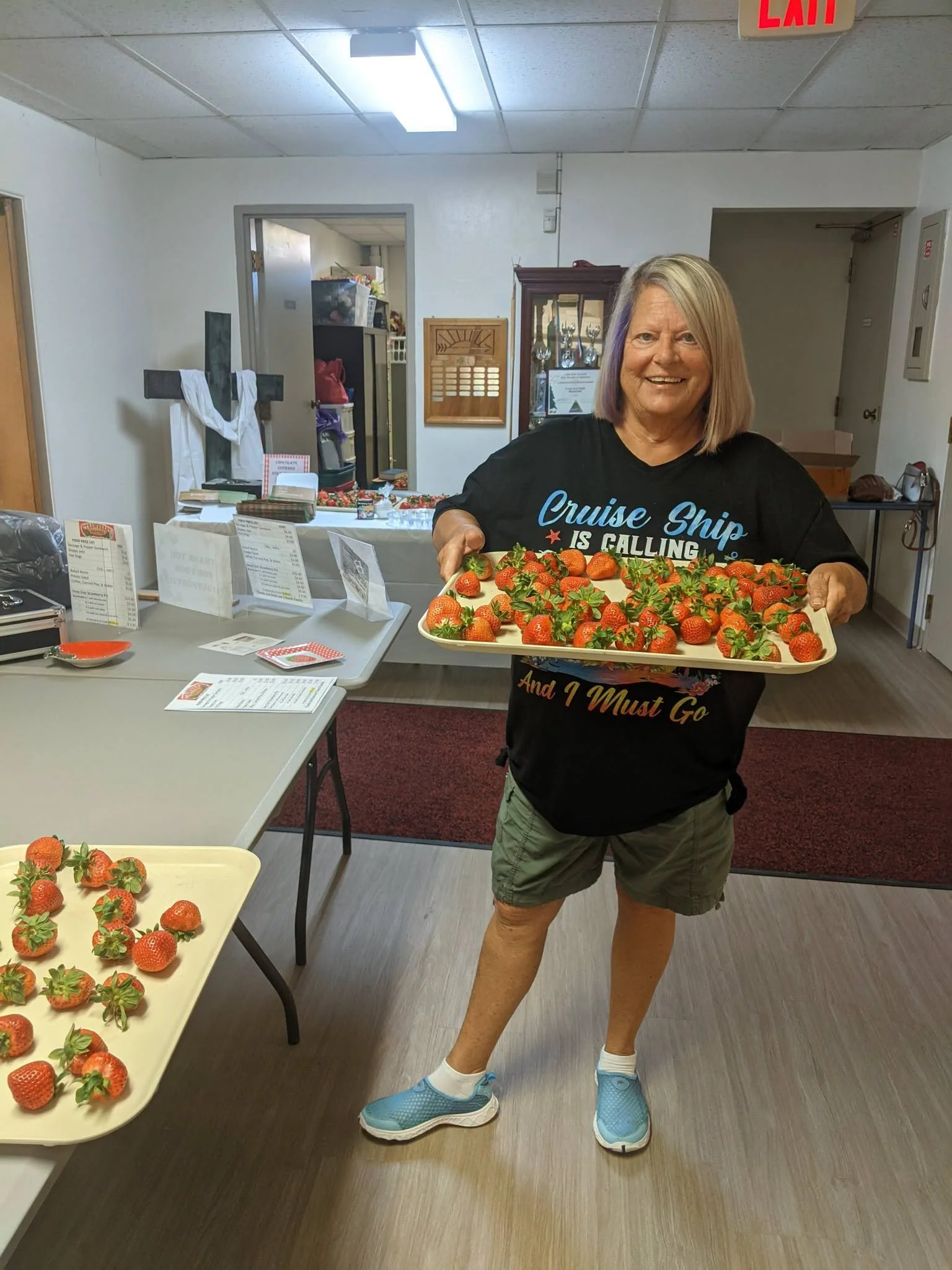 A woman smiling and holding a tray of strawberries inside a room, with tables and checkout counters visible in the background.