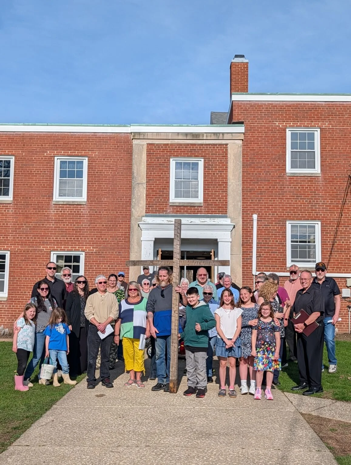 Group of people gathered in front of a brick building holding a large wooden cross, celebrating a religious or community event.
