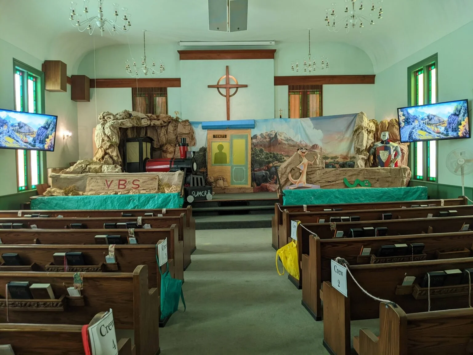 View of a church stage decorated with cartoon theme, including a train, a character from Ice Age, and a knight armor, with two television screens displaying scenic images on either side, and wooden pews in the foreground.