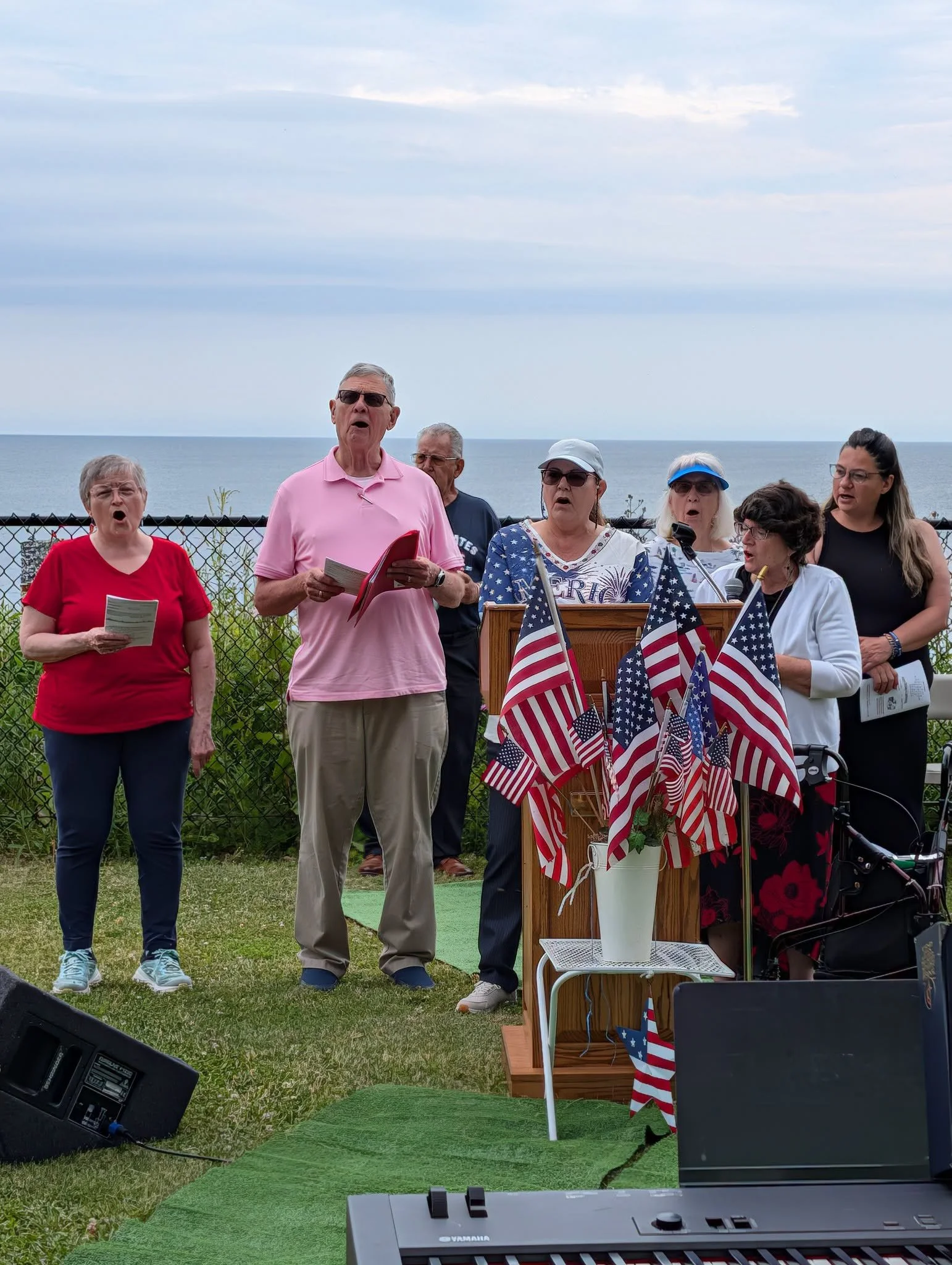 A group of six older adults performing a song outdoors near the ocean, with American flags and patriotic decorations on a wooden stand.