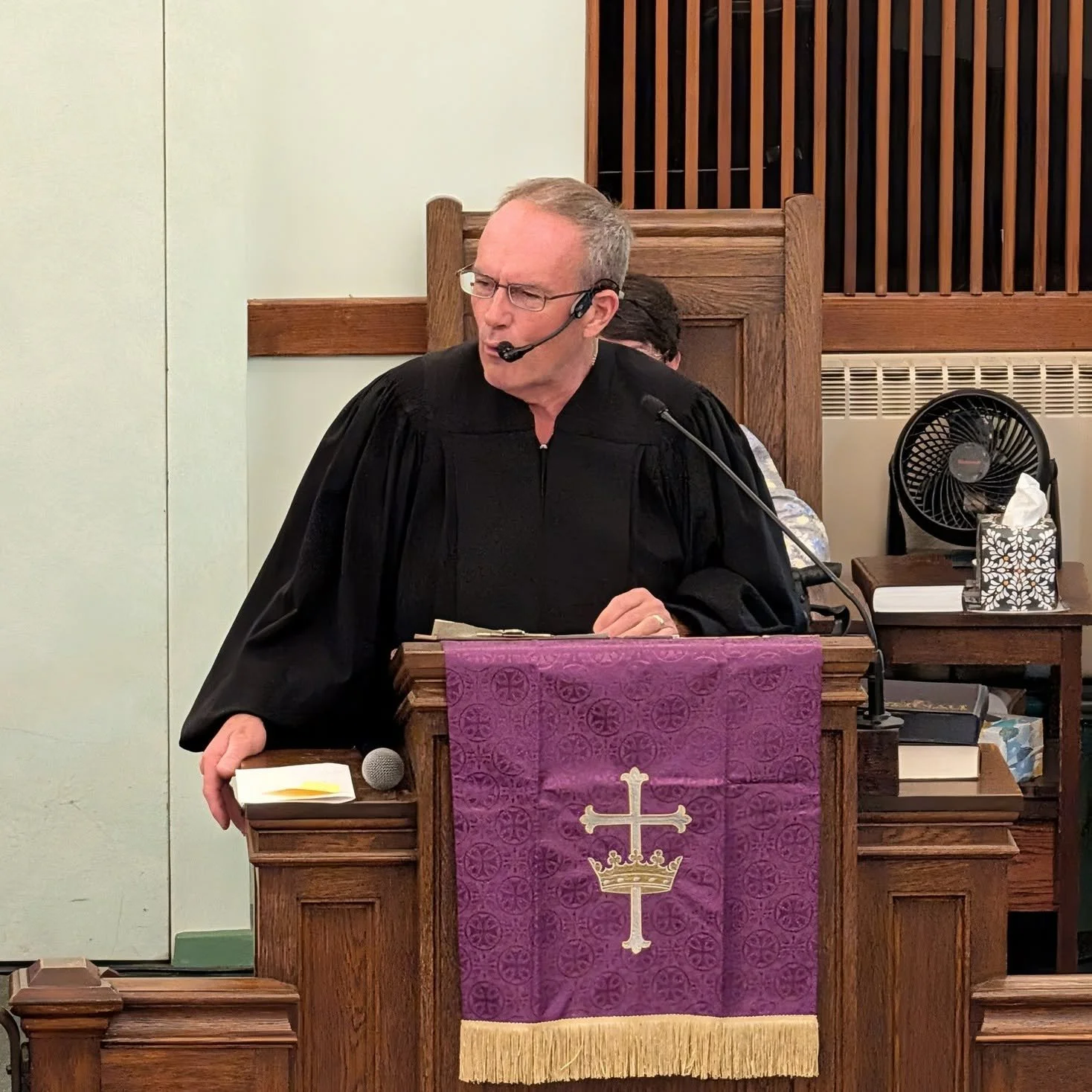 A man in a black robe and glasses stands at a wooden pulpit in a church, speaking into a headset microphone, with a purple cloth featuring a cross and crown hanging from the pulpit.