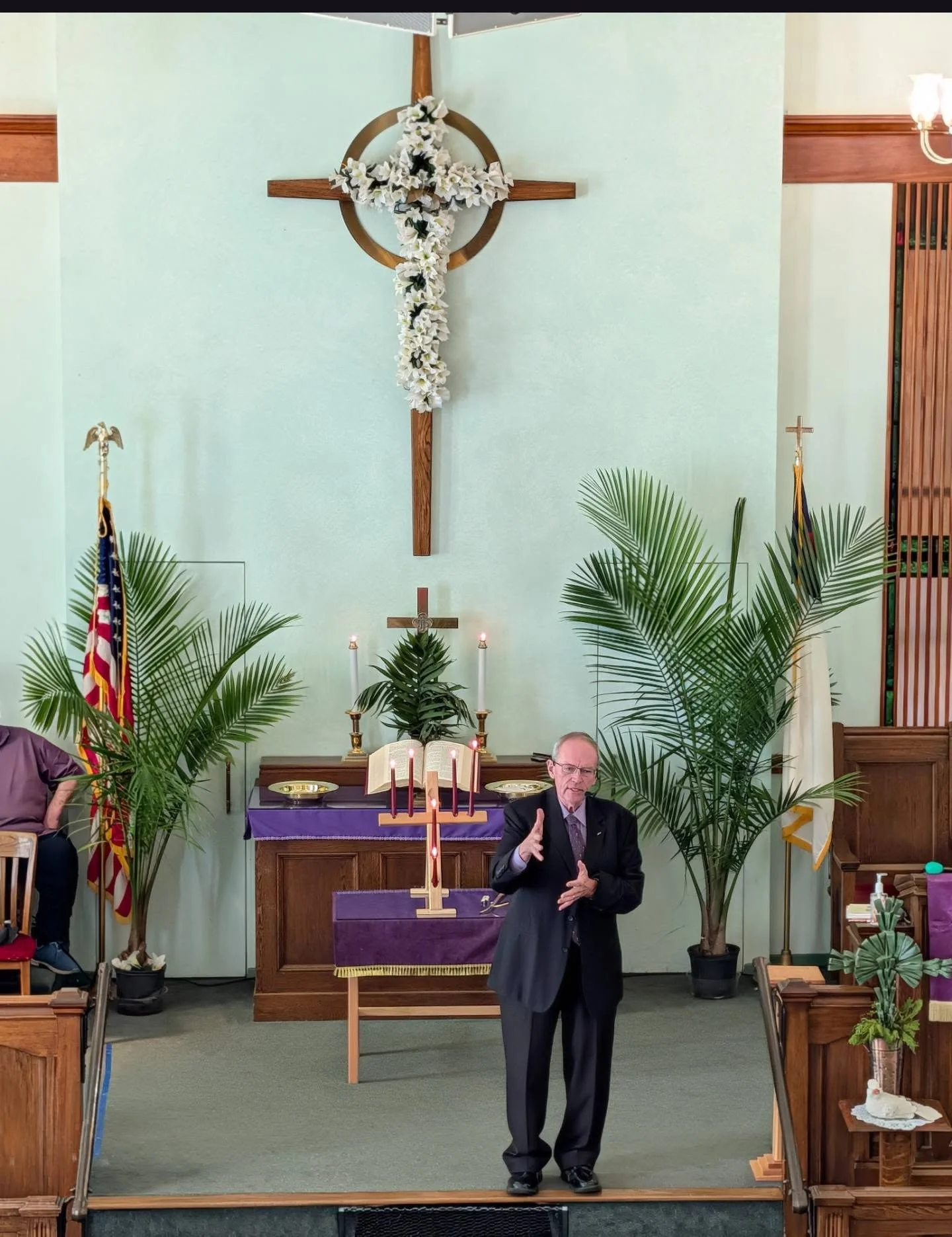 A man in a suit and glasses is speaking or giving a sermon in a church with a cross on the wall behind him. The church is decorated with large green plants, candles, and religious symbols, including an open Bible on a purple cloth on a stand.