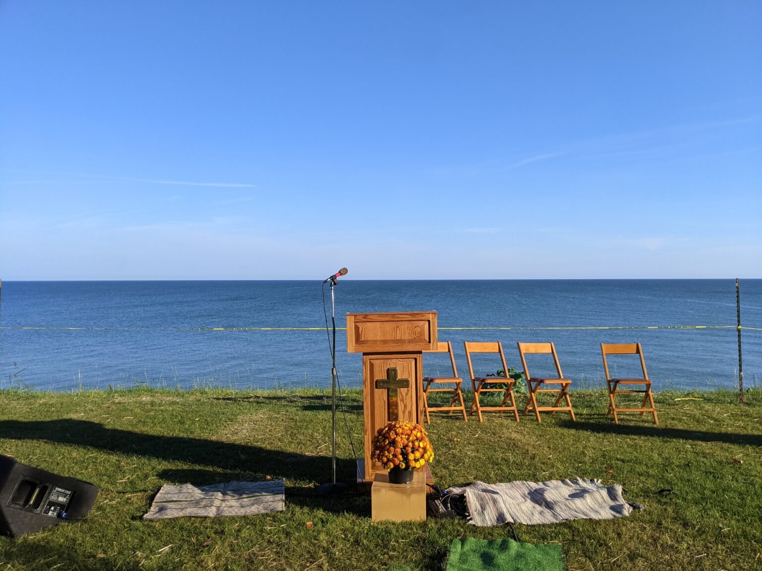 Outdoor setup with a wooden pulpit featuring a cross, a flower pot, and four yellow chairs facing a large body of water under a clear blue sky, prepared for a ceremony or event.