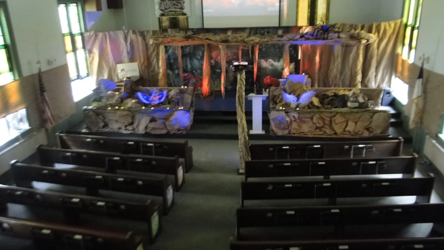 Empty church sanctuary with benches and a stage decorated with a wilderness theme, including rocks, trees, and animal figures, lit with blue and red lights.