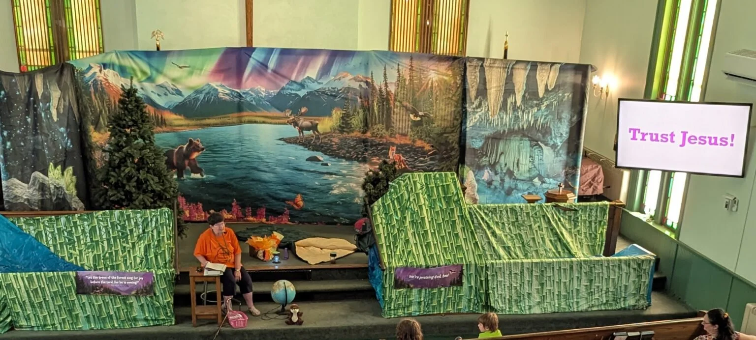A stage with a biblical scene backdrop of a river, mountains, and wilderness, decorated with trees, animals, and nature elements. People are seated, with one woman sitting on a stool near a globe, in front of a screen displaying 'Trust Jesus!'