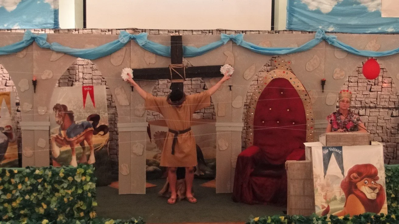 A theatrical performance background with stone castle walls, blue drapes, and fantasy-themed posters. A performer is in the center dressed as a monk, holding pink and white pom-poms, with balloons and a woman sitting at a desk on the right side.